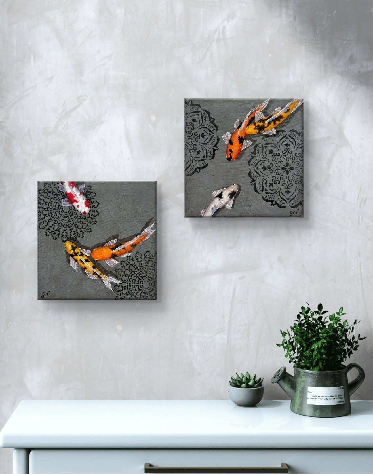 Two square artworks of koi fish on a gray wall above a white cabinet with plants.